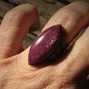 May include: A marquise-shaped ring featuring a polished, deep magenta stone set in a dark, oxidized metal band. The stone has a smooth, slightly textured surface with subtle variations in color, securely held within a simple metal bezel setting.