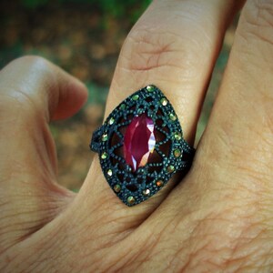 May include: A dark, ornate ring featuring a large, marquise-cut ruby center stone. The ring has a black metal setting with intricate filigree and small, sparkling accents. The ring is shown on a hand.