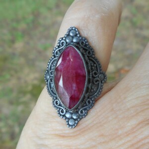 May include: A marquise-cut deep red gemstone ring set in an oxidized silver band. The metal setting features intricate scrollwork and small bead details surrounding the central stone, creating a pointed, elongated shape on the finger.