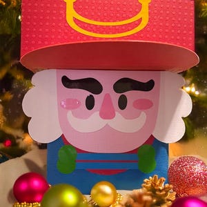 Peut inclure: Une boîte-cadeau en carton rouge et bleu en forme de casse-noisette avec un visage blanc, des yeux noirs, un nez rose et une moustache. La boîte est entourée d'ornements de Noël dorés, verts et roses.