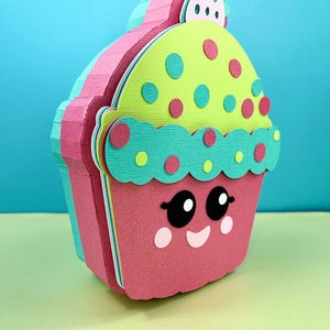 May include: A pink, green, and blue cupcake with a smiling face and white polka dots. The cupcake has a green frosting with red and yellow polka dots and a white cherry on top.