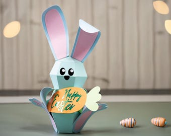 Puede incluir: Un conejito de papel de color azul claro sosteniendo un cartel con forma de zanahoria que dice "Feliz Pascua". El conejito tiene orejas rosas y ojos negros. Hay dos pequeñas zanahorias rayadas de color naranja y blanco sobre la mesa.