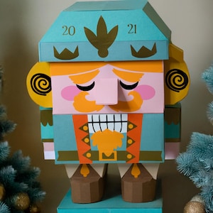 Può includere: Una grande figura di schiaccianoci di carta colorata con un corpo e una testa blu-verde, capelli e baffi arancioni e un viso rosa. La figura ha i numeri "20" e "21" sulla testa. Decorazioni per l'albero di Natale sullo sfondo.