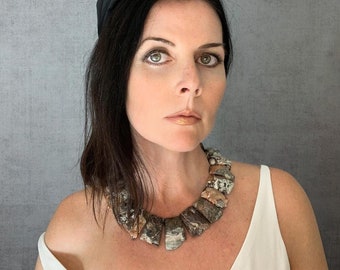 May include: A woman with dark hair is wearing a black headband and a statement necklace made of large, irregularly shaped, multi-colored stones. The necklace is a chunky, geometric design.