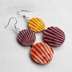 May include: A pair of earrings with three round, textured shell-shaped beads. The beads are yellow, purple, and orange. The earrings have silver hooks.