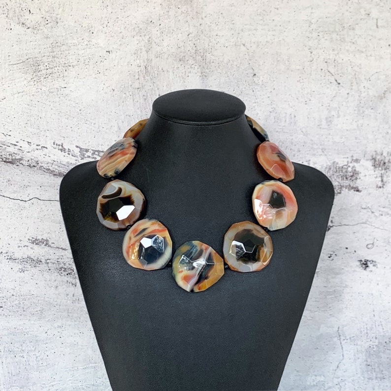 May include: A statement necklace featuring large, faceted, circular stones in shades of orange, brown, black, and cream. The stones are arranged in a single row, showcasing a polished, multi-colored banded pattern with a reflective, geometric cut surface.