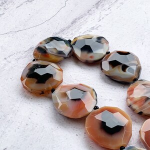 May include: A strand of faceted, polished agate beads featuring a marbled pattern of translucent orange, deep black, and creamy white tones. Each bead has a rounded, irregular shape with multiple flat facets that reflect light across the surface.