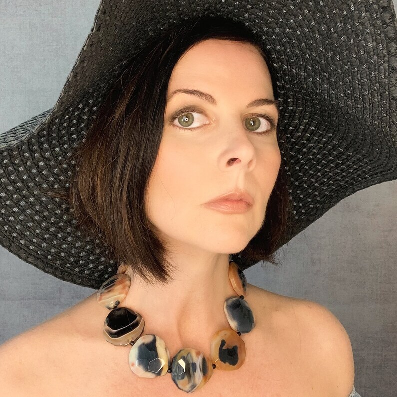 May include: A statement necklace featuring large, polished, faceted agate stones in shades of translucent amber, cream, and deep black. The stones are strung together with small black beads, creating a bold, multi-colored accessory that rests against the collarbone.