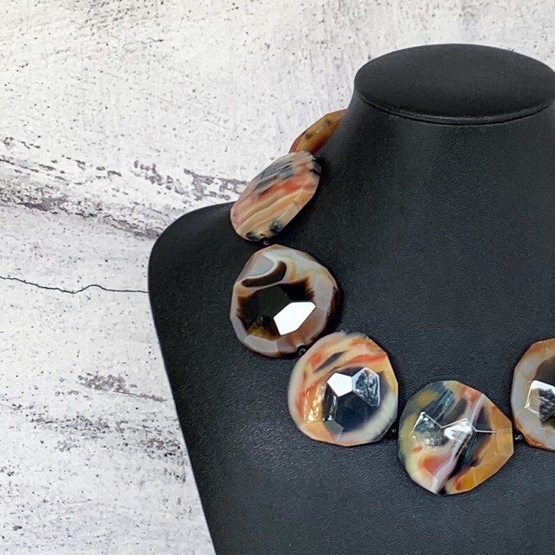 May include: A statement necklace featuring large, faceted, irregular-shaped agate stones. The stones display a natural banded pattern with warm tones of orange, brown, cream, and black, showcasing a polished, reflective surface on a black display bust.