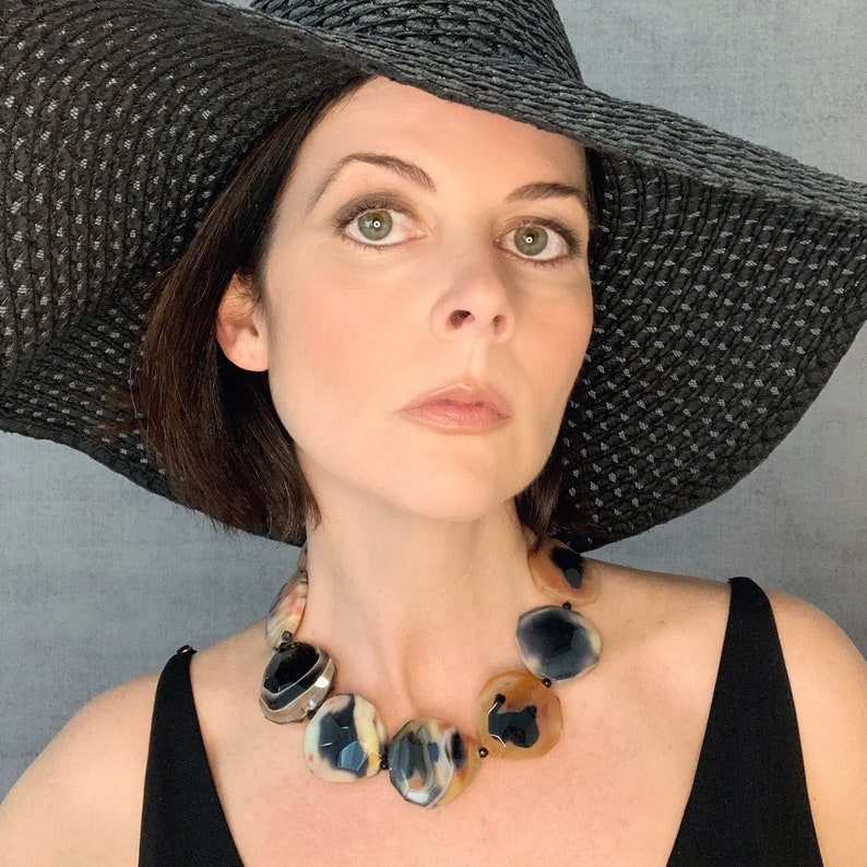 May include: A statement necklace featuring large, polished, multi-faceted stones in shades of translucent amber, cream, and deep black. The stones are linked together in a circular arrangement, creating a bold, chunky design that rests against the neck.