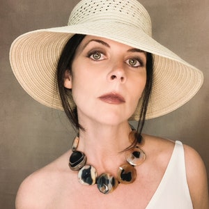 May include: A wide-brimmed beige straw hat paired with a statement necklace featuring large, polished, multi-colored agate stones in shades of black, cream, and brown, strung together on a dark cord, worn with a simple white sleeveless top.