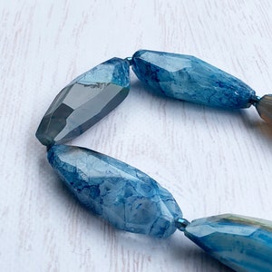 May include: Close-up of a necklace featuring large, faceted beads. The beads are primarily shades of blue, with some appearing translucent and others with a marbled effect. The beads are strung together with a thin, blue string, creating a striking jewelry piece.