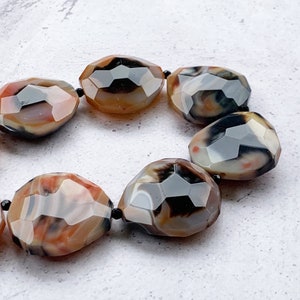 May include: A bracelet composed of faceted, irregular, oval-shaped agate beads. The beads feature a marbled pattern with earthy tones of brown, cream, black, and hints of orange, separated by small, round, black spacer beads on a string.