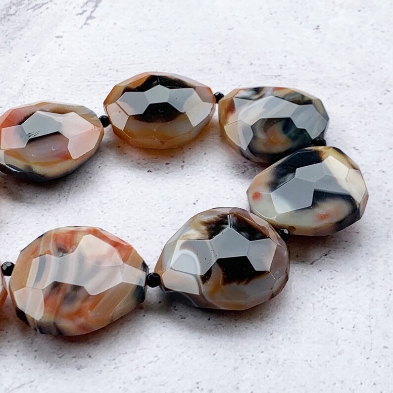 May include: A bracelet composed of faceted, irregular, oval-shaped agate beads. The beads feature a marbled pattern with earthy tones of brown, cream, black, and hints of orange, separated by small, round, black spacer beads on a string.