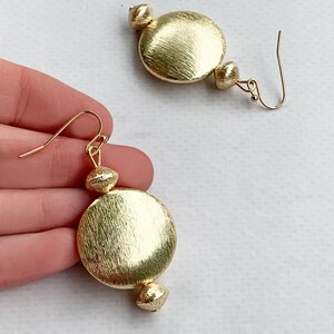 Könnte beinhalten: Goldfarbene Ohrringe mit strukturiertem, gebürstetem Finish. Jeder Ohrring hat eine große, runde Scheibe, eine kleinere Perle und einen Hakenverschluss. Die Ohrringe werden vor einem weißen Hintergrund präsentiert.