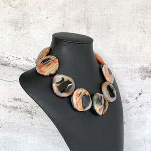 May include: A statement necklace featuring large, faceted, polished agate stones in shades of orange, cream, black, and gray. The stones are arranged in a circular pattern, showcasing natural banding and translucent areas within each individual piece.
