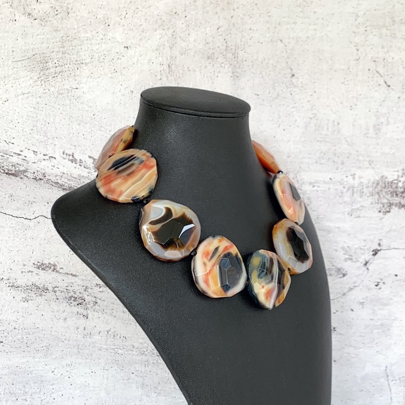 May include: A statement necklace featuring large, faceted, polished agate stones in shades of orange, cream, black, and gray. The stones are arranged in a circular pattern, showcasing natural banding and translucent areas within each individual piece.