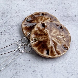 May include: A pair of brown and white ceramic earrings with a textured surface. The earrings are attached to silver hooks.