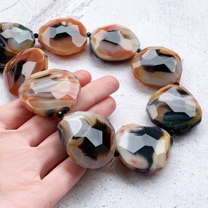 May include: A necklace featuring large, faceted, irregular-shaped beads in shades of translucent orange, cream, and deep black. The polished stones are strung together with small black spacer beads, creating a bold, multi-colored statement piece.