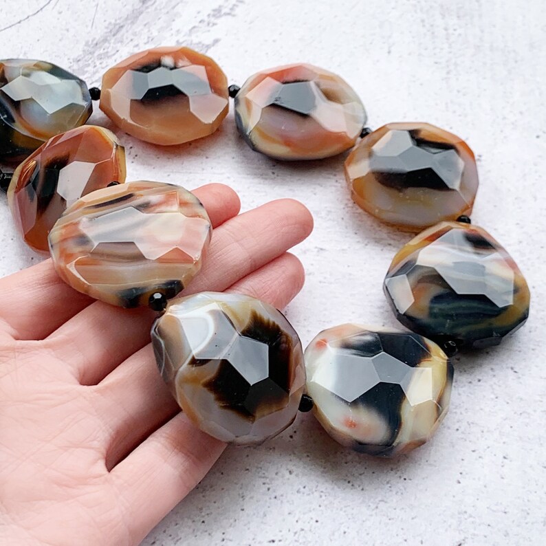 May include: A necklace featuring large, faceted, irregular-shaped beads in shades of translucent orange, cream, and deep black. The polished stones are strung together with small black spacer beads, creating a bold, multi-colored statement piece.
