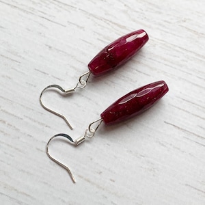 May include: A pair of drop earrings with faceted, oblong, deep red gemstones. The gemstones are suspended from silver-coloured fish hook ear wires. The earrings are displayed on a light-coloured, textured surface.
