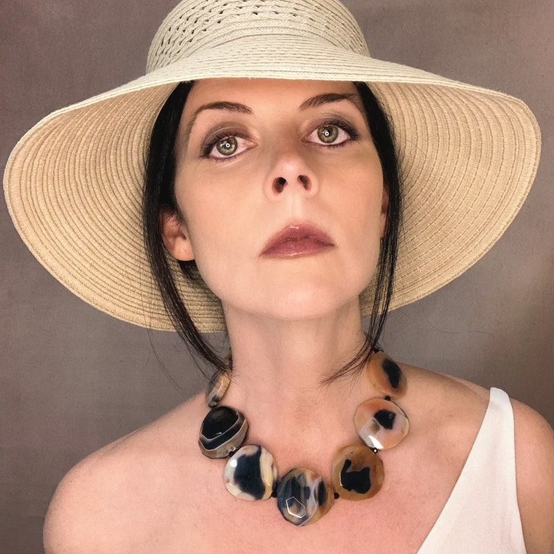 May include: A statement necklace featuring large, polished, circular agate stones in shades of black, cream, and translucent amber. The stones are strung together on a dark cord, creating a bold, multi-colored accessory worn around the neck.
