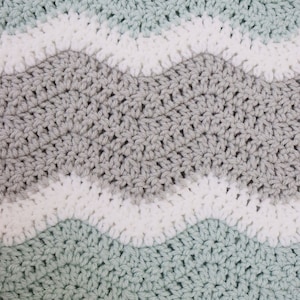 May include: A crocheted blanket featuring a wavy pattern in shades of light green, grey, and white. The blanket is crafted with a textured stitch and has a soft, cosy feel.
