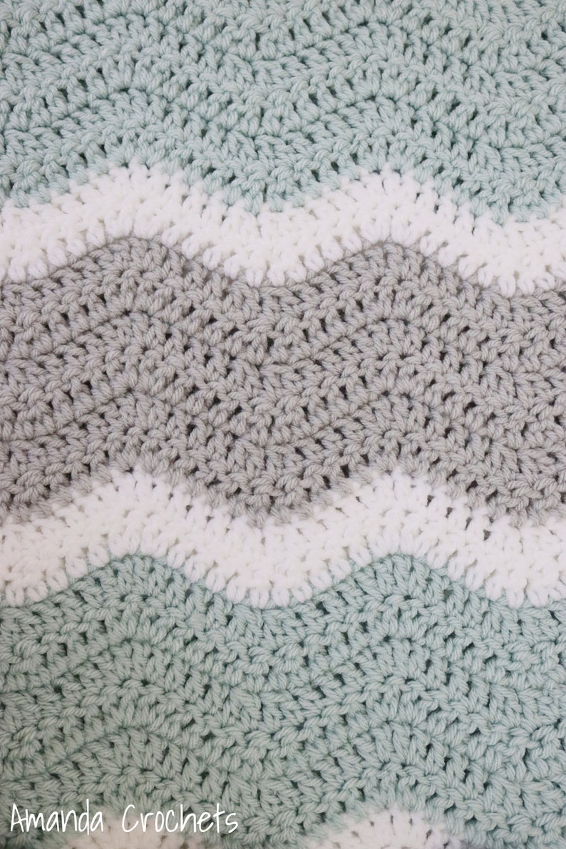 May include: A crocheted blanket featuring a wavy pattern in shades of light green, grey, and white. The blanket is crafted with a textured stitch and has a soft, cosy feel.