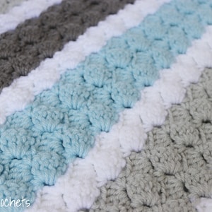Pode incluir: Uma manta de crochê listrada com fileiras alternadas de branco, azul claro, cinza e branco. A manta é feita com um ponto texturizado que cria um padrão em relevo.