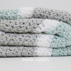 May include: A crocheted baby blanket in shades of grey, white, and light blue. The blanket is folded in thirds and is made with a simple stitch pattern.