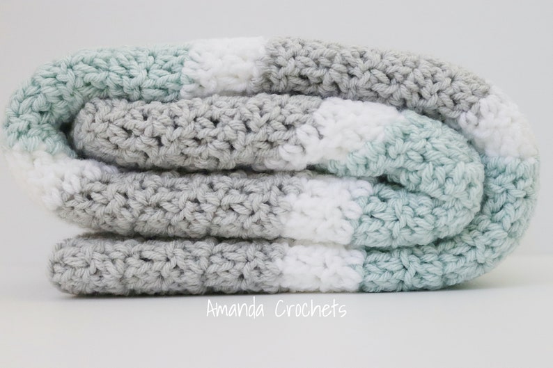 May include: A crocheted baby blanket in shades of grey, white, and light blue. The blanket is folded in thirds and is made with a simple stitch pattern.