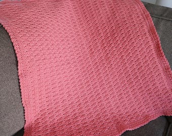 May include: A coral-colored, crocheted blanket with a textured pattern. The blanket is square-shaped and has a decorative border. The blanket is folded over a gray sofa.
