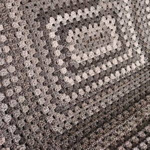 May include: A handmade crochet blanket in shades of gray, white, and black. The blanket features a granny square design, with a central square and radiating rows of squares. The texture is soft and inviting, perfect for home decor.