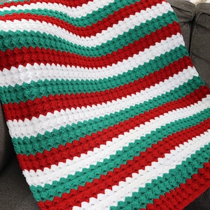 May include: A handmade crochet blanket in a striped pattern of red, white, and green. The blanket is folded over a gray sofa, showcasing the textured stitch and vibrant colors. This cozy blanket is perfect for home decor.