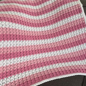 Könnte beinhalten: Eine gestrickte Babydecke in rosa und weiß mit Streifen. Die Decke ist mit einem strukturierten Stich gearbeitet und hat einen weißen Rand.