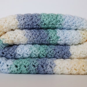 May include: A crocheted baby blanket in shades of blue, green, and cream. The blanket is folded in thirds and is made with a textured stitch.