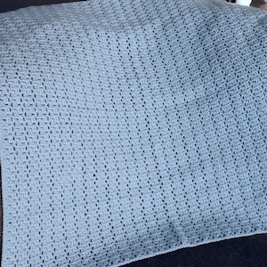 May include: A light blue crocheted blanket with a textured pattern. The blanket is rectangular and appears to be handmade. The blanket is resting on a brown surface.