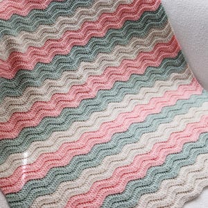 May include: A crochet blanket featuring a repeating ripple pattern in alternating horizontal stripes of soft pink, sage green, and cream. The textile displays a consistent wavy texture throughout, created by the interlocking yarn stitches in these colors.
