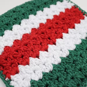 May include: A square, handmade crochet item in a granny square pattern. The color scheme features alternating stripes of green, white, and red. The stitches are visible, creating a textured surface. The item is likely a decorative piece or a small blanket.