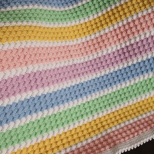 May include: A handmade, colorful crochet blanket featuring alternating stripes of pastel pink, purple, blue, green, and yellow. The blanket has a textured stitch pattern and a white scalloped border. A cozy, soft blanket.