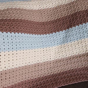 May include: A handmade crochet blanket with horizontal stripes in shades of brown, cream, and blue. The blanket is draped over a brown surface, showcasing the soft texture and color variations. The blanket is approximately 50 inches wide.