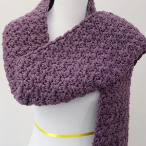 May include: A purple crocheted scarf on a white mannequin. The scarf is long and wide, and it is made with a simple stitch.