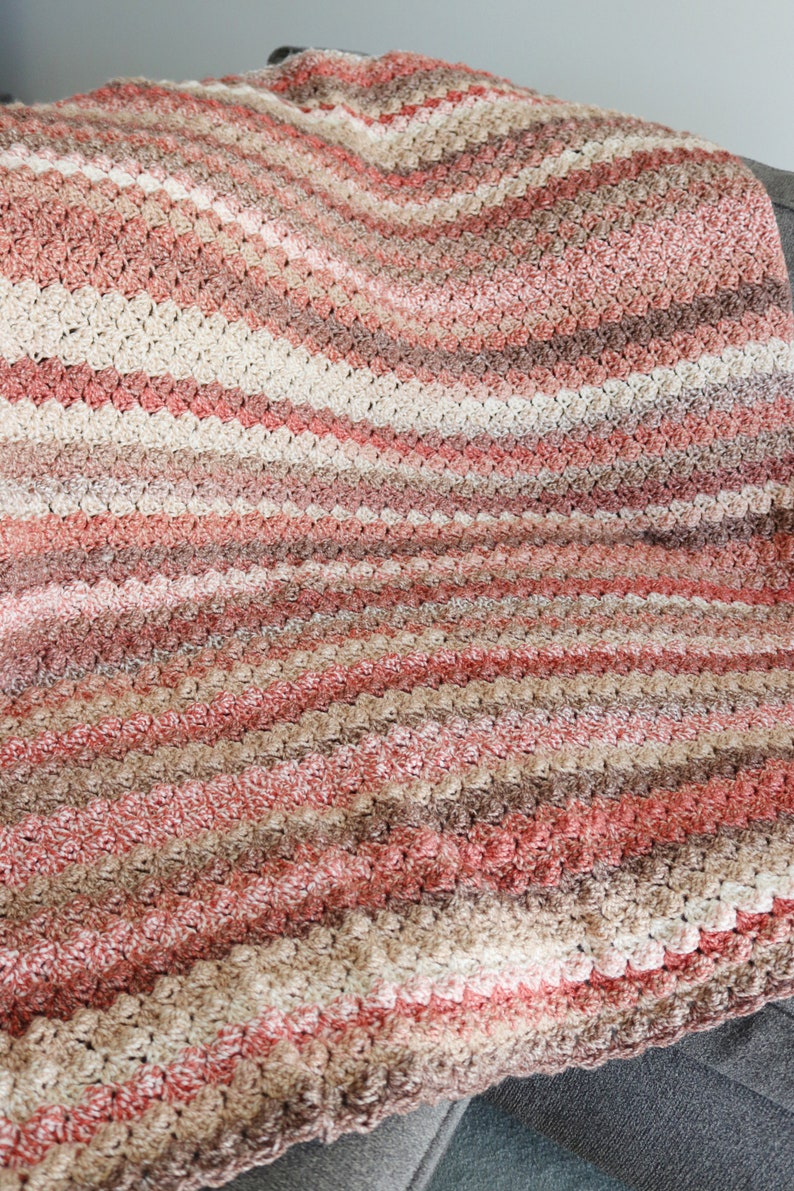 K&ouml;nnte beinhalten: Eine geh&auml;kelte Decke mit einem gestreiften Muster in Rosat&ouml;nen, Braun und Beige. Die Decke ist mit einem strukturierten Stich gearbeitet, der eine erhabene, unebene Oberfl&auml;che erzeugt.