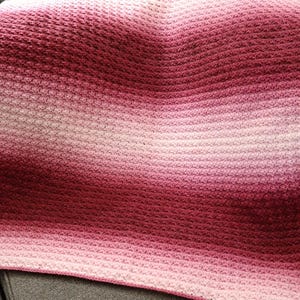 May include: A crocheted blanket with a gradient of pink and red colors. The blanket is made with a textured stitch and has a soft, fuzzy appearance.