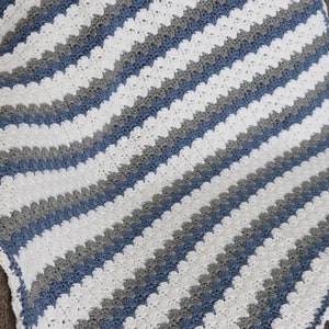 Pode incluir: Um cobertor de bebé em crochê com um padrão chevron em branco, cinza e azul.