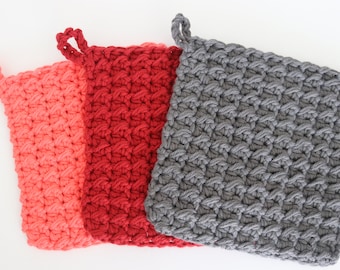 Puede incluir: Tres agarraderas de olla de ganchillo en coral, rojo y gris. Las agarraderas de olla son cuadradas con un asa de bucle en la parte superior.