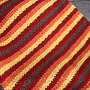 Pode incluir: Uma manta de crochê com listras diagonais em tons de vermelho, laranja, amarelo e marrom.