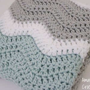 May include: A crocheted baby blanket in shades of grey, white, and light blue. The blanket is made with a wavy stitch pattern.