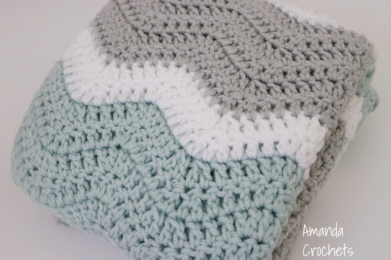 May include: A crocheted baby blanket in shades of grey, white, and light blue. The blanket is made with a wavy stitch pattern.