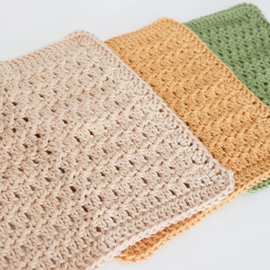 Peut inclure: Trois torchons en crochet de différentes couleurs : beige, orange et vert. Les torchons sont empilés les uns sur les autres, le beige étant en haut.